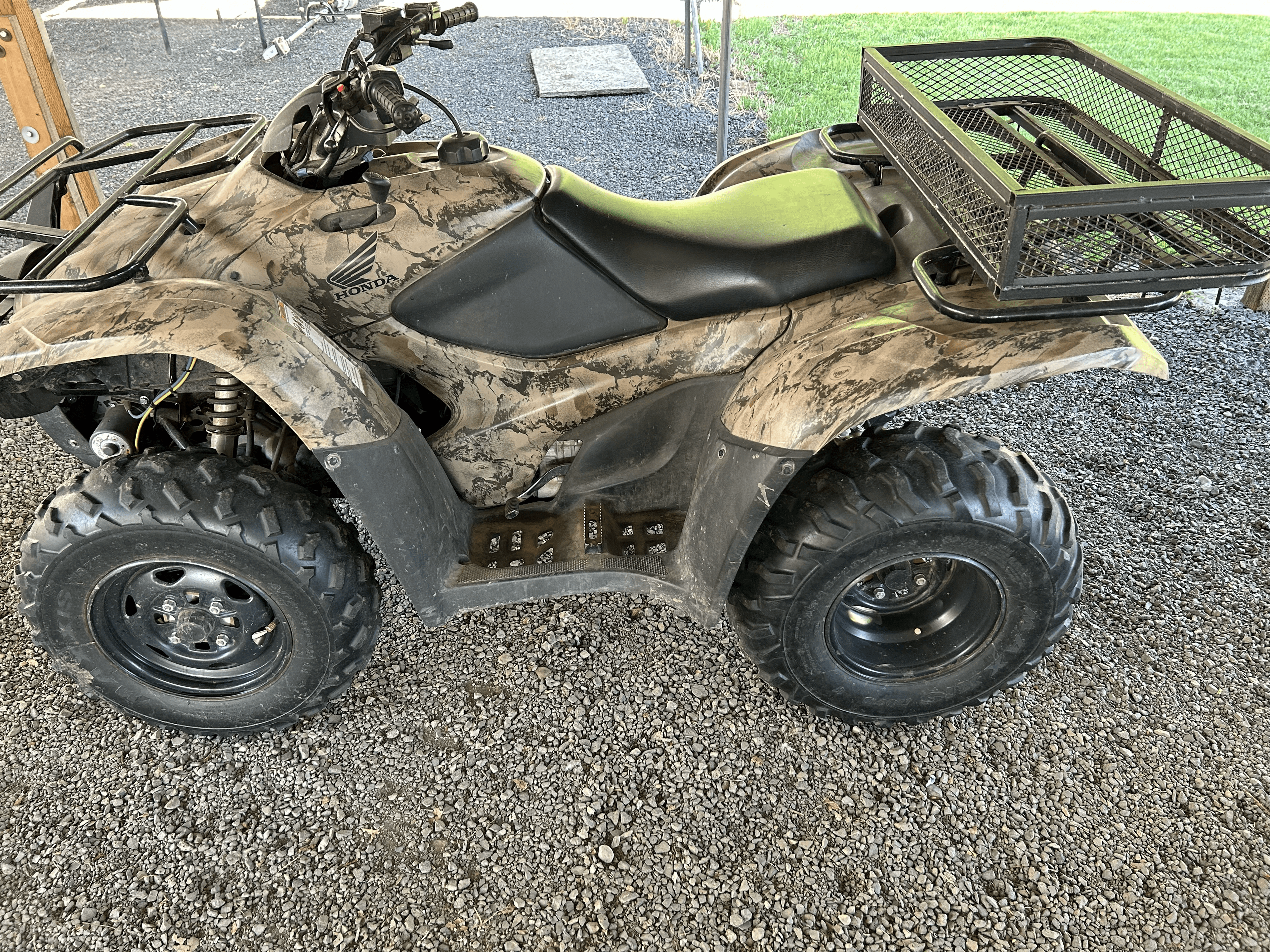 Camouflage Honda ATV with a black seat and rear metal cargo basket on gravel.